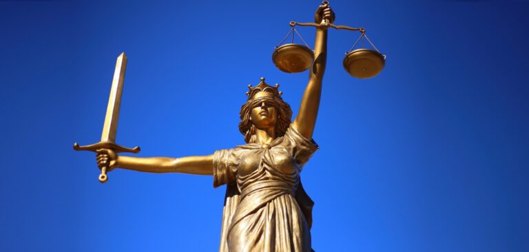 Χρυσαφί αγαλματίδιο της Δίκης, της θεάς της δικαιοσύνης, με το πρόσωπό της καλυμμένο. Κρατάει ένα σπαθί στο αριστερό της χέρι και μία ζυγαριά στο δεξί. Η θεά Δίκη φοράει ένα στέμμα στο κεφάλι της. Ο ουρανός στο παρασκήνιο είναι καθαρός και μπλε,